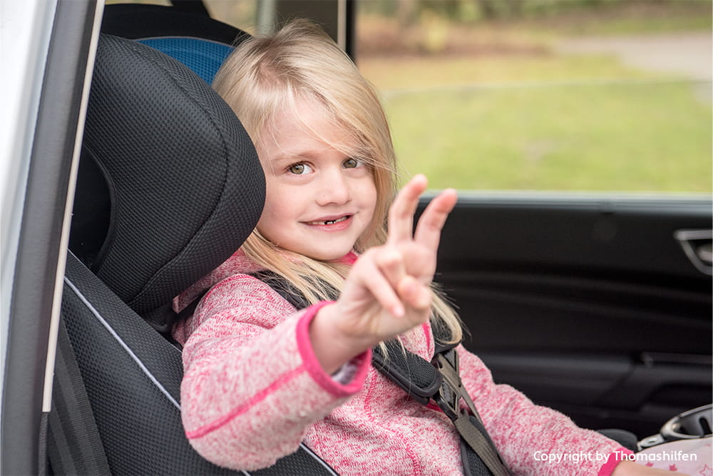 Kind sitzt angeschnallt im Auto-Kindersitz und zeigt ein Peace-Zeichen