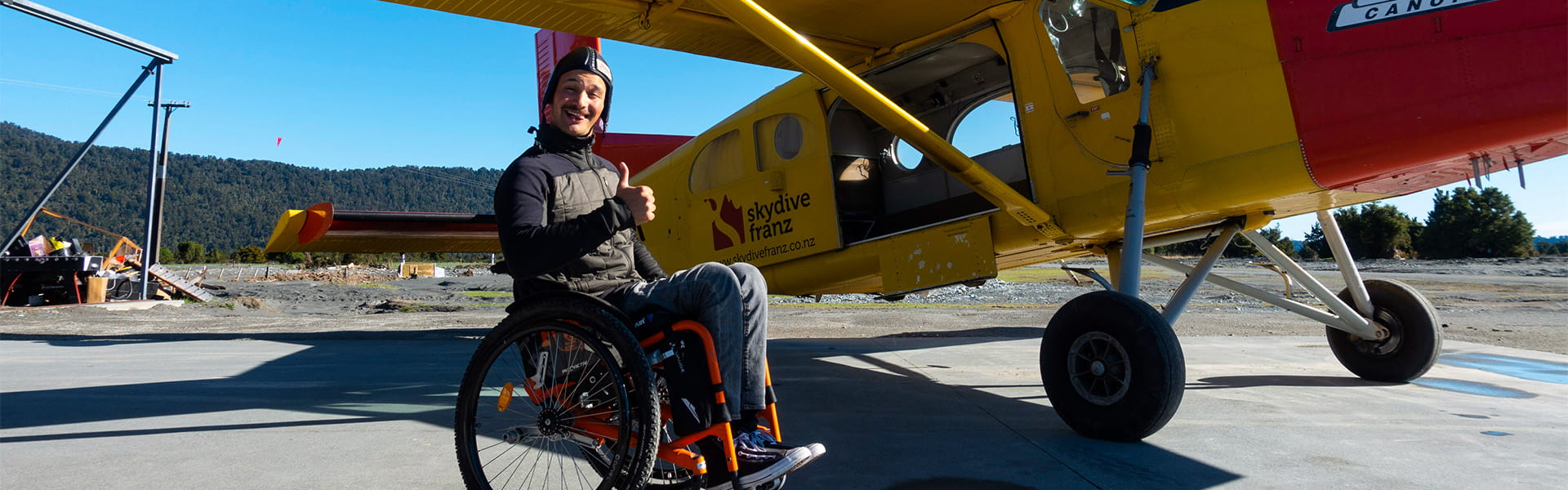 Ein Mann im Rollstuhl steht vor einem kleinen Flugzeug auf dem Flugplatz und zeigt einen Daumen hoch.