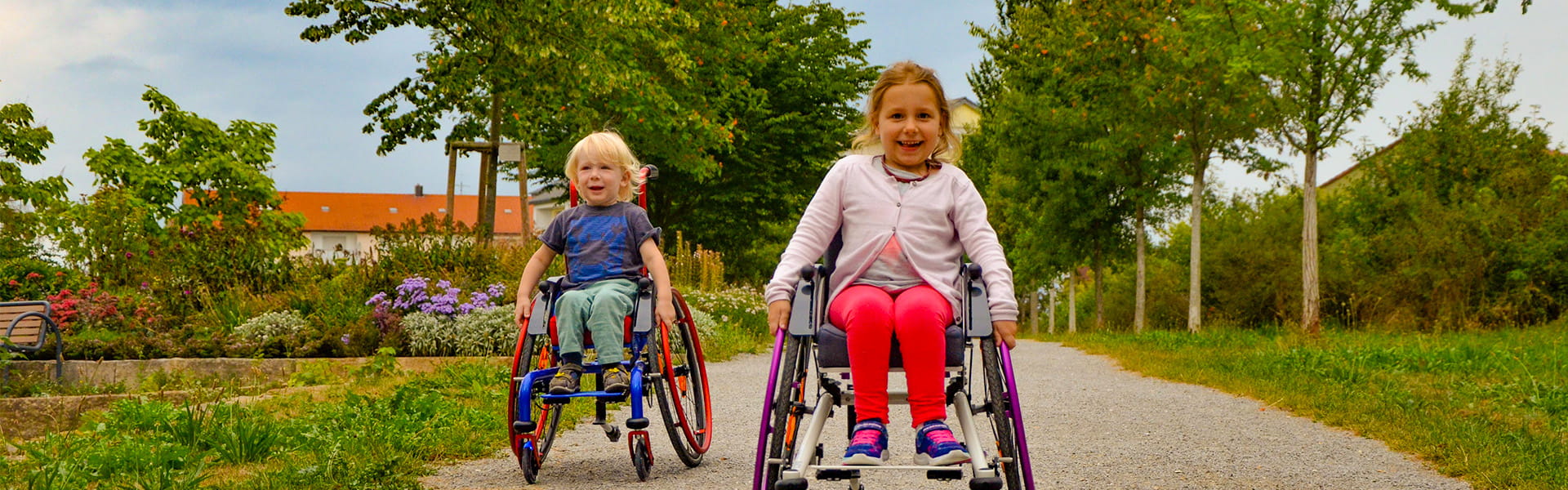 Zwei Kinder im Rollstuhl fahren im Park auf die Kamera zu.