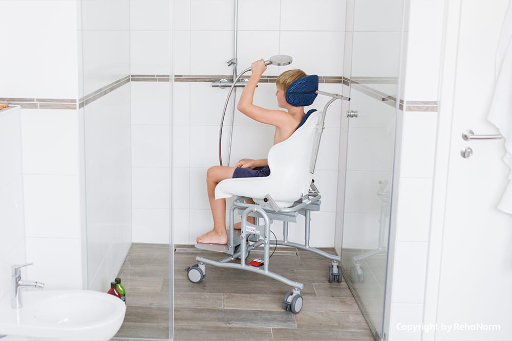 Kind sitzt auf einer mobilen Duschhilfe in einer barrierearmen Dusche und hält die Duschbrause