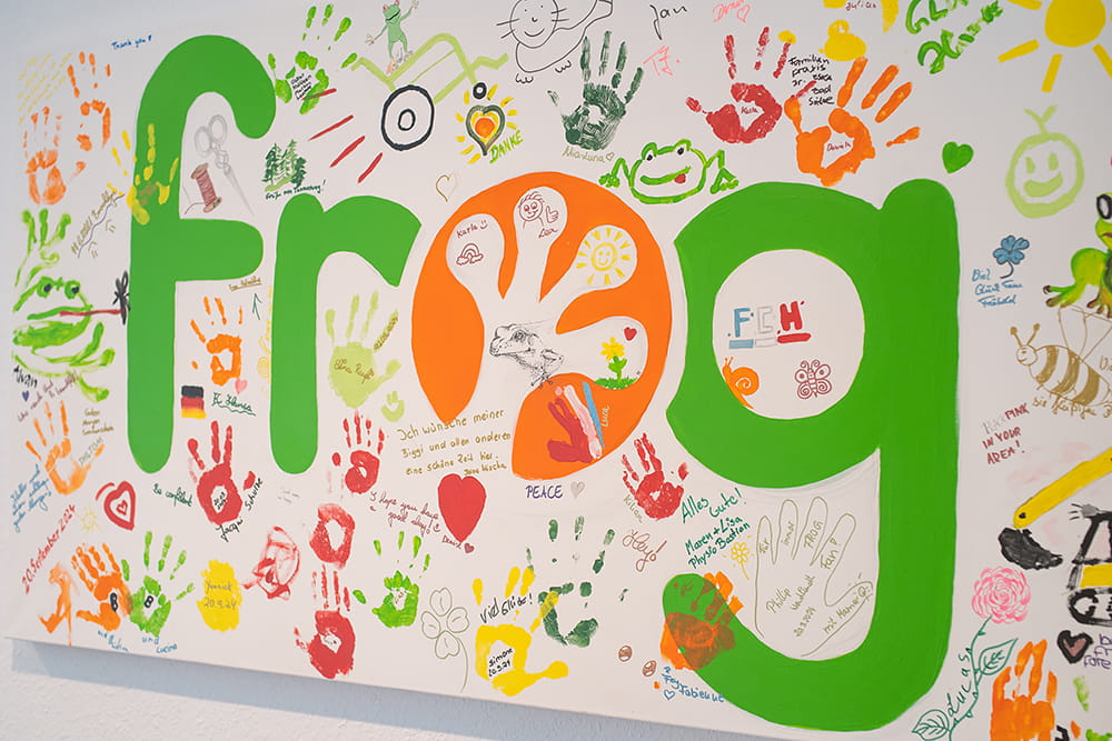Bunte Wand mit FROG-Logo, Handabdrücken, Zeichnungen und persönlichen Grüßen von Kindern