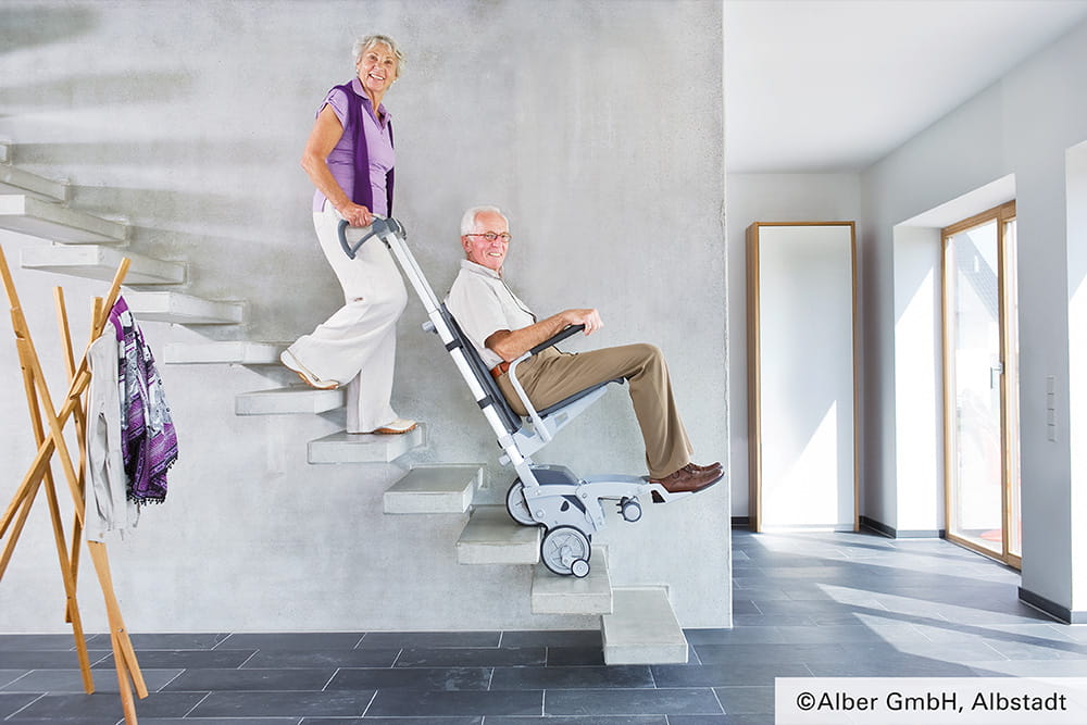 Eine Frau transportiert ihren Mann mit einem Treppenlifter die Treppe herunter.