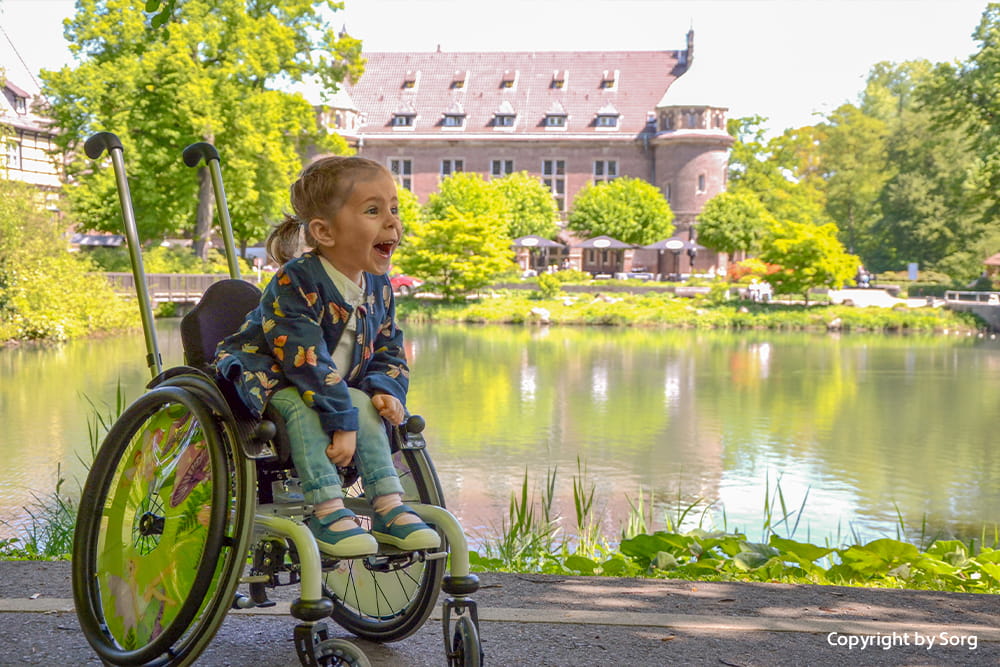 Lachendes Kind sitzt in einem farbenfrohen Rollstuhl am See vor einer grünen Parklandschaft