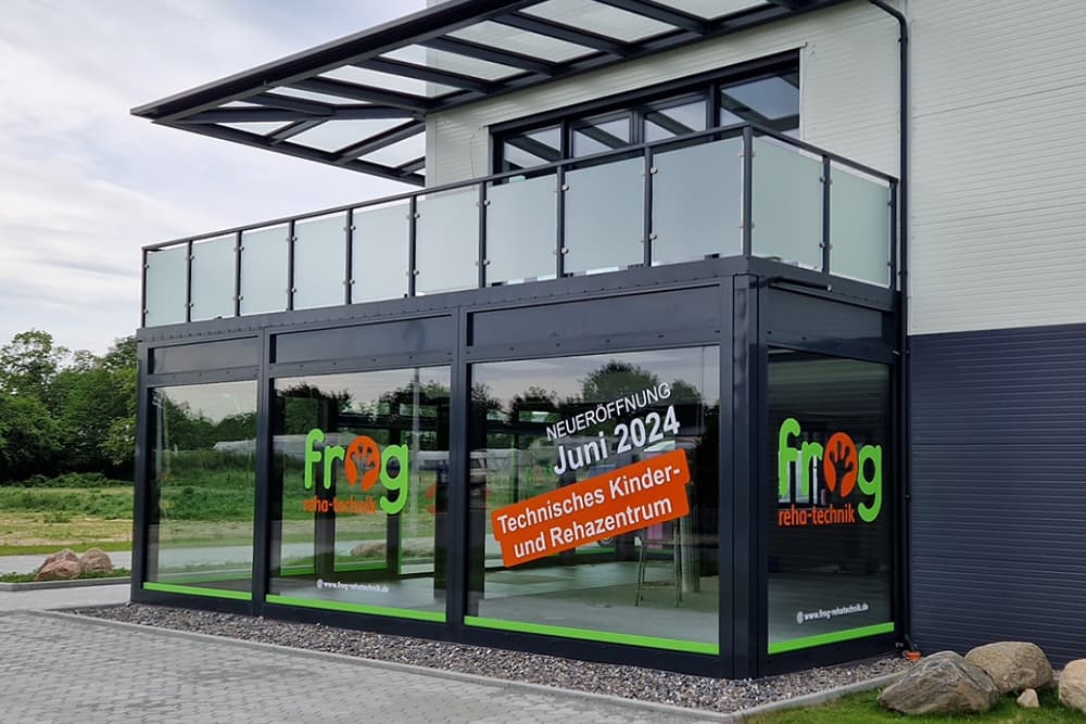 Eine Hausecke mit gläsernem Vorbau. Auf der Fensterfront sind Aufkleber mit dem Logo der FROG und eine Information über die Neueröffnung.
