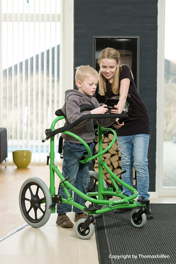 Kind steht in einem grünen Gehtrainer, während ein älteres Kind daneben gemeinsam auf ein Gerät schaut