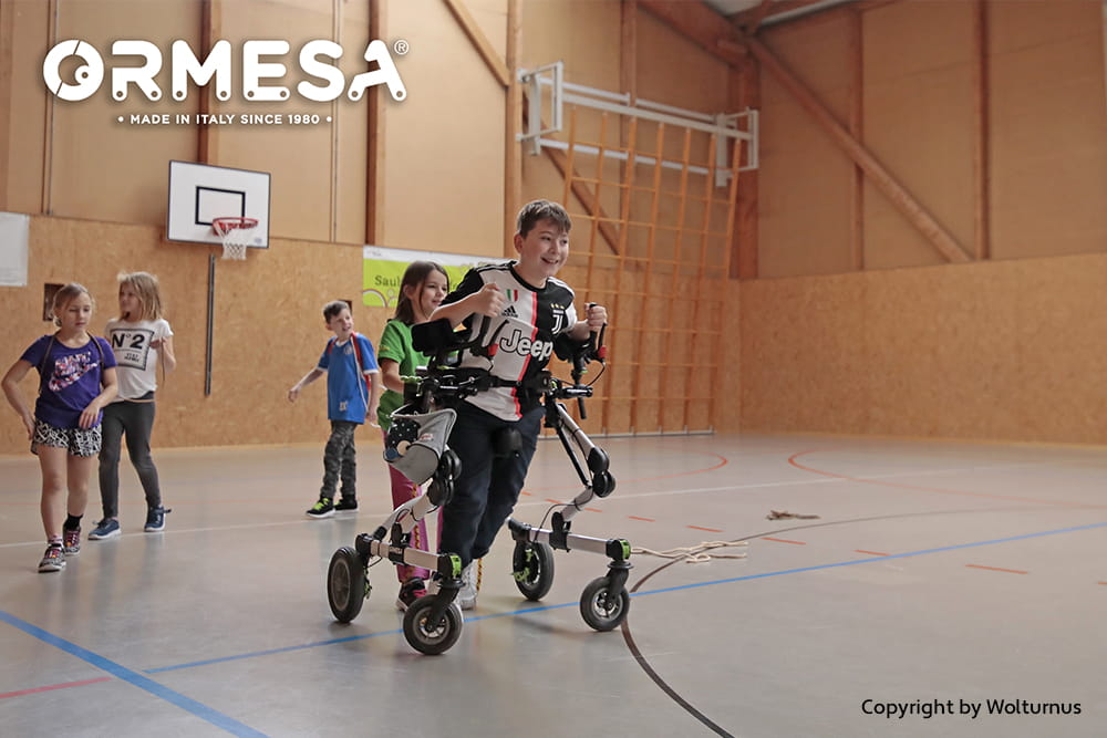 Kind nutzt einen Gehtrainer in einer Sporthalle und nimmt gemeinsam mit anderen Kindern an einer Aktivität teil