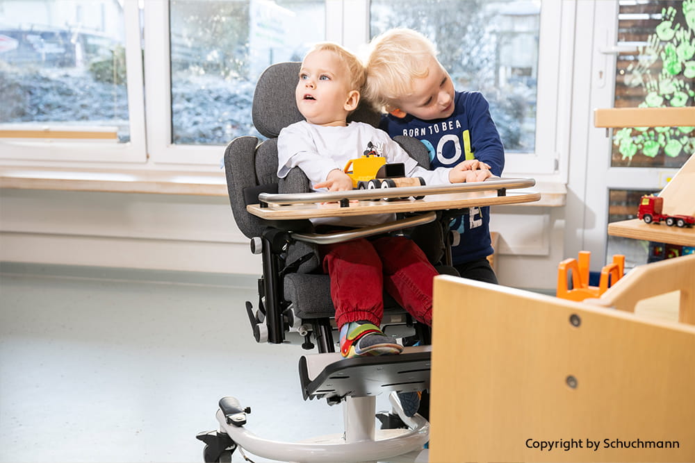 Kind sitzt in einem Therapiestuhl mit Tischplatte, während ein anderes Kind daneben spielt