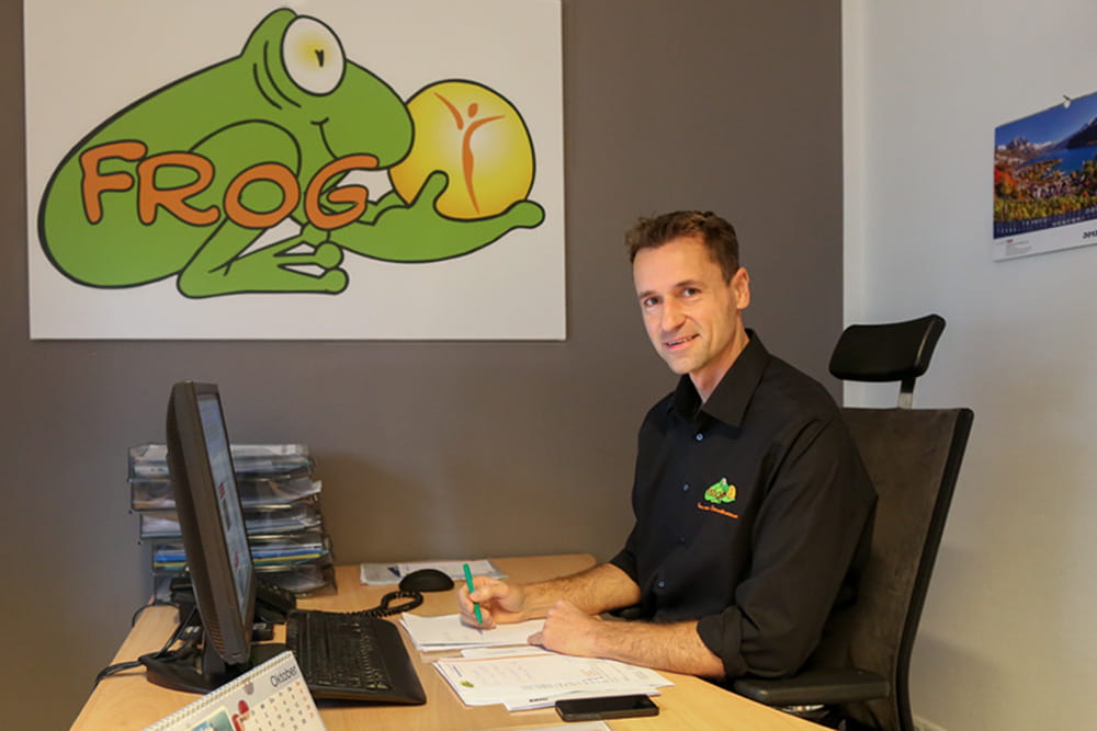 Jan Wüstenberg sitzt im Büro und lächelt in die Kamera. An der Wand ist der FROG Frosch groß zu sehen.