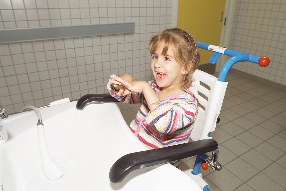 Ein kleines Mädchen sitzt in einem speziellen Rollstuhl am Waschbecken und freut sich.
