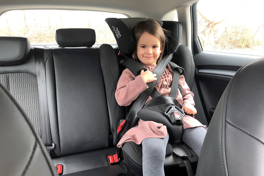 Ein kleines Mädchen sitzt im Auto-Kindersitz. Sie lächelt dabei und hält sich etwas am Gurt fest.
