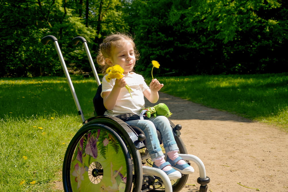 Ein kleines Mädchen im Rollstuhl sitzt im Park und spielt mit Löwenzahn.
