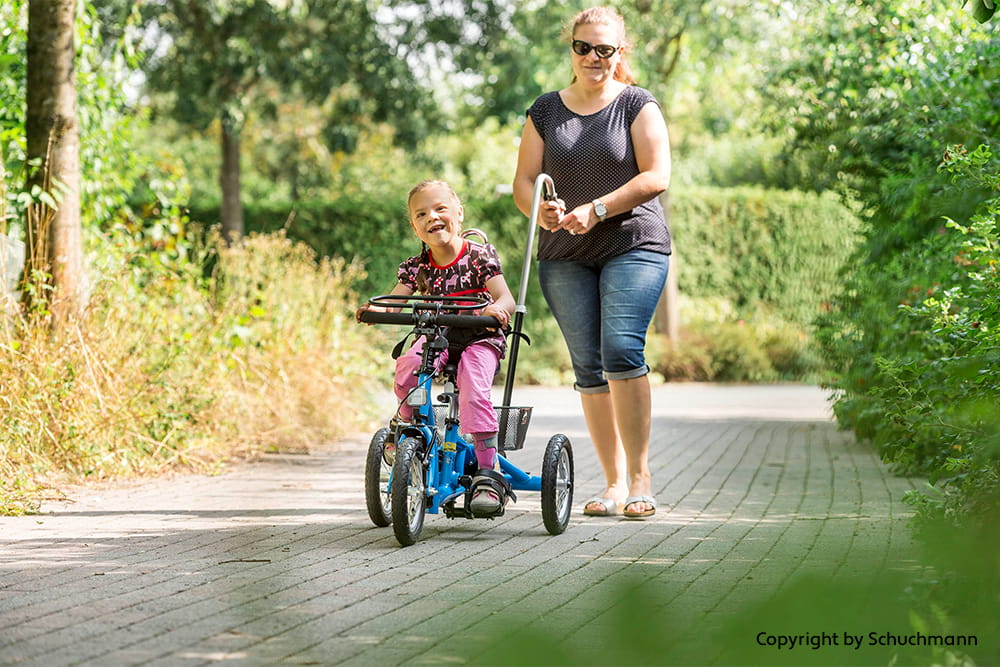 Kind fährt ein blaues Therapiedreirad, während eine Begleitperson das Dreirad mit einer Stange unterstützt