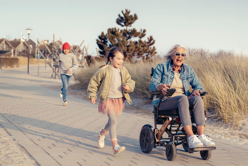 Oma fährt mit dem elektrischen Rollstuhl auf einem Pflasterweg und ein kleines Mädchen läuft lachend daneben.