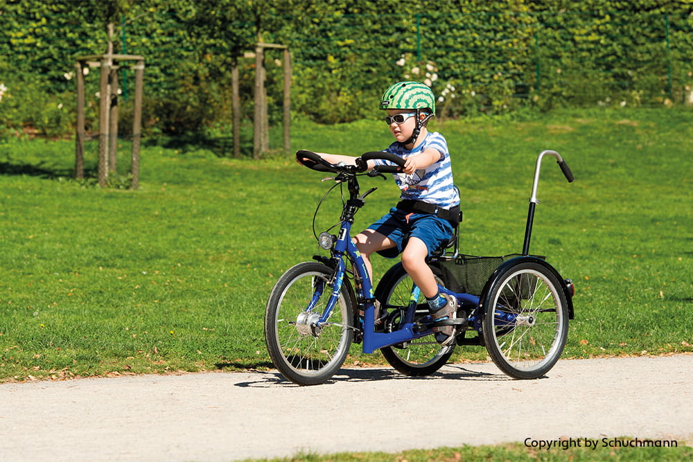 Kind fährt mit Helm auf einem blauen Therapiedreirad durch einen Park