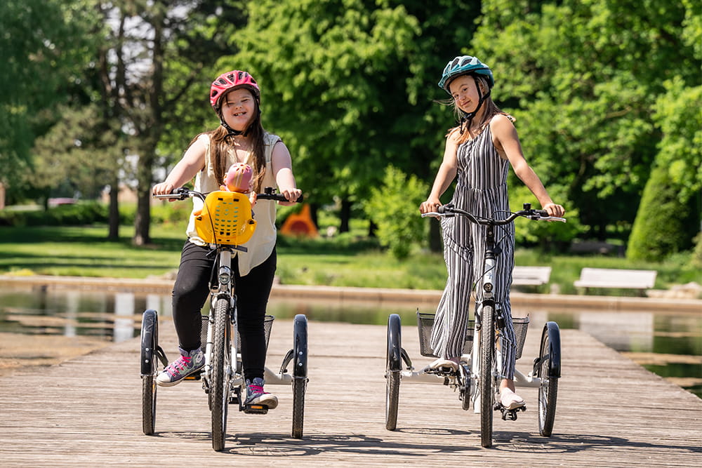 Zwei mädchen fahren auf Reha Fahrrädern in einem Park im grünen auf einem Weg. Beide tragen Schutzhelme.