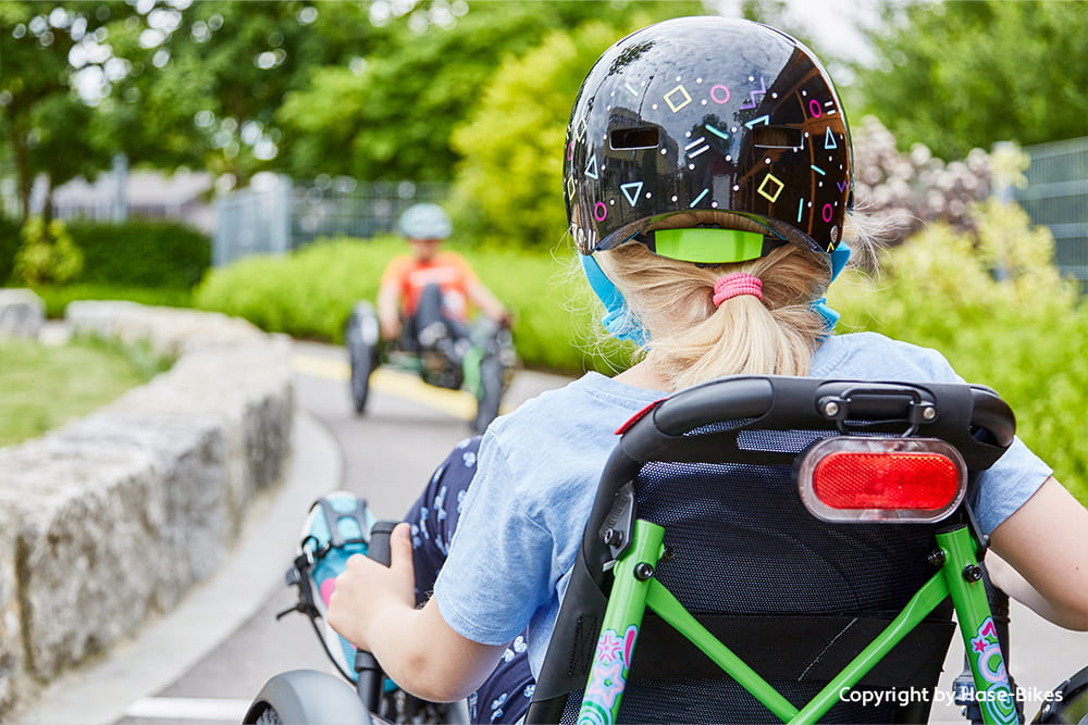 Kind fährt mit Helm auf einem grünen Therapie-Dreirad hinter einer weiteren Person auf einem Liegerad