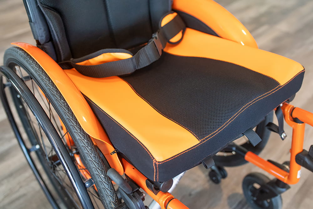 Nahaufnahme einer orange-schwarzen Sitzfläche eines Rollstuhls mit Gurt und stabiler Polsterung
