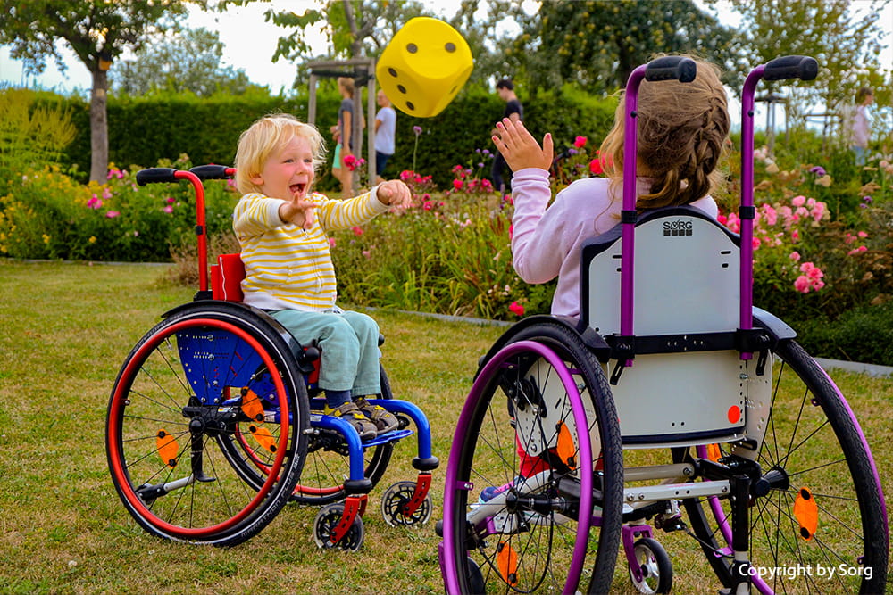 Zwei Kinder in bunten Rollstühlen spielen im Park mit einem gelben Würfelball