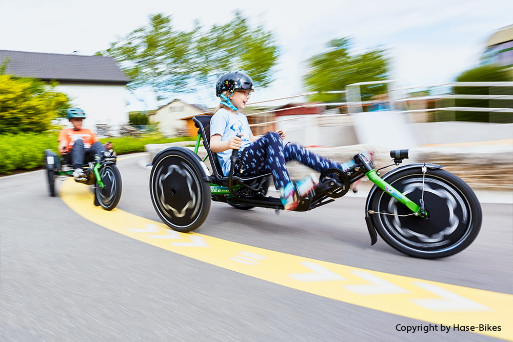Zwei Kinder fahren mit Helm auf einem grünen Therapie-Dreirad die Straße entlang
