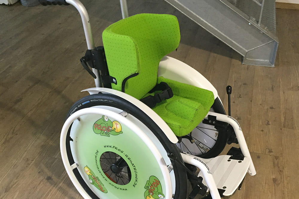 Ein umgebauter Rollstuhl der FROG. Die Sitzfläche ist grün und die Reifenschützer sind mit dem FROG Logo versehen.